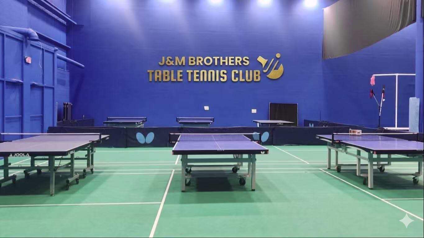 J&M Brothers Table Tennis Club interior — professional tables on green courts beneath the club logo on the blue wall